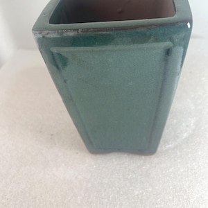 May include: A square-shaped ceramic planter with a glossy dark green glaze. The vessel features a tapered design with a recessed panel on each side and a smooth, open top. The interior reveals a natural terracotta-colored clay finish.