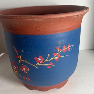 May include: A terracotta and blue ceramic planter. The top and base are terracotta, while the body is blue with a painted floral design featuring red flowers and gold branches. The planter has a rounded shape.