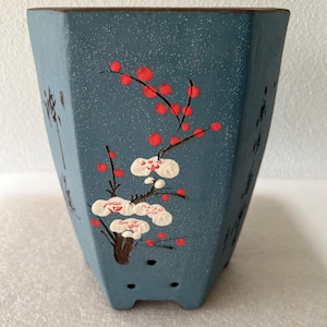 May include: A teal-colored, octagonal ceramic planter with a textured finish. The planter features a hand-painted design of red berries and white blossoms on dark branches. The sides have Asian-style calligraphy.