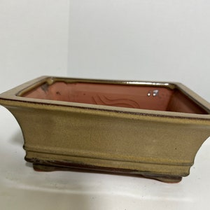 May include: A square, light brown ceramic bonsai pot with a slightly raised rim and a textured surface. The pot has a dark brown glaze around the rim and base.