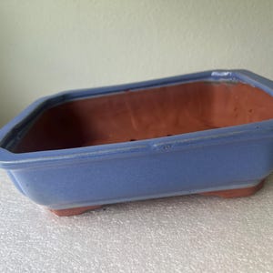 May include: A rectangular bonsai pot with a smooth periwinkle blue glazed exterior and a natural terracotta interior. The vessel features a wide rim, slightly tapered sides, and four small feet at the base, with drainage holes visible inside.