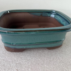 May include: A square bonsai pot with a teal glaze and a brown interior. The pot has a rectangular shape with rounded corners and sits on small feet. The rim has a glossy finish, and the interior is a contrasting brown color.