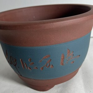 May include: A terracotta bonsai pot with a blue band and a brown inscription. The inscription is in Chinese characters.