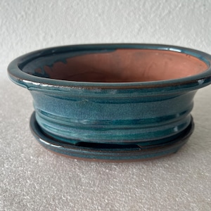 May include: A teal-glazed ceramic bonsai pot with a matching saucer. The pot has a wide, shallow design with a slightly flared rim and a textured surface. The interior is a warm terracotta color, suitable for small plants.