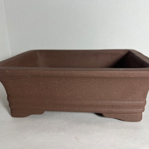 May include: A brown, rectangular bonsai pot with a textured surface. The pot has a square shape with slightly flared sides and small feet. The pot is designed for growing bonsai trees.