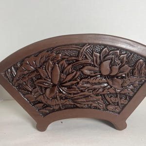 May include: A brown ceramic planter shaped like a fan with a raised design of lotus flowers and leaves.