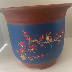 May include: A terracotta flower pot with a blue glaze and a painted design of a bird on a branch with red berries. The pot has a rounded bottom and a wide rim.