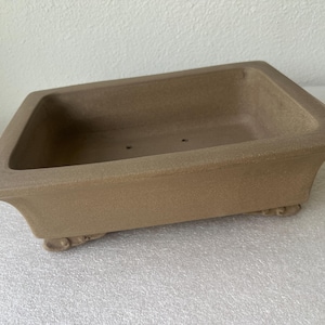May include: A rectangular bonsai pot in a light brown color. The pot has a textured surface and a slightly flared rim. It features decorative feet and drainage holes, suitable for bonsai trees or other small plants.