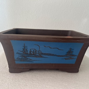 May include: A rectangular brown ceramic bonsai pot featuring a wide, flat rim and a flared base. The front side displays a rectangular blue panel decorated with a dark, etched landscape illustration of trees and a shoreline scene.