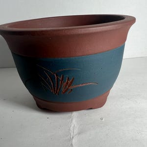 May include: A ceramic planter with a two-tone design. The top and bottom are a reddish-brown, while the middle section is a teal blue. A decorative plant design is etched into the blue section.