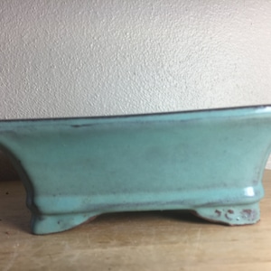 May include: A light blue rectangular ceramic bonsai pot with four feet. The pot has a slightly textured surface.