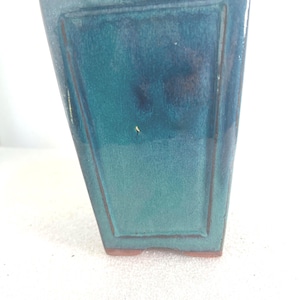 May include: A rectangular ceramic planter with a glossy teal and blue glaze. The vessel features a tapered shape with a raised border frame on the front panel and a visible terracotta-colored base, showcasing a smooth, reflective surface finish.