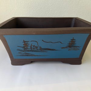 May include: A square bonsai pot with a brown rim and base, and a blue center panel. The blue panel features a brown illustration of a landscape with a pagoda and other elements. The pot has a slightly flared design.