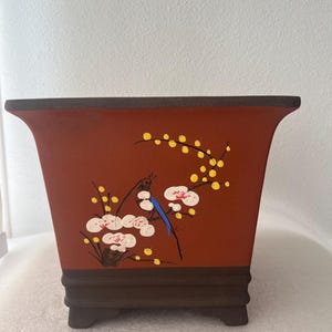May include: A square terracotta-colored ceramic planter with a dark brown base and four small feet. The front features a hand-painted design of a small bird with a blue wing perched on a branch with white blossoms and small yellow berries.