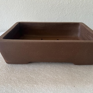 May include: A brown, rectangular bonsai pot with a slightly textured surface. The pot has a square shape with angled sides and small feet. The interior of the pot is visible, and it has drainage holes. The pot is designed for bonsai trees.