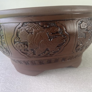 May include: A brown ceramic bonsai pot with a smooth rim and a rounded body. The exterior features embossed panels depicting a bird in flight surrounded by clouds and floral motifs. The vessel stands on four small, integrated feet at the base.