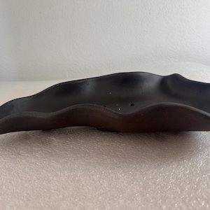 May include: A dark brown, leaf-shaped ceramic dish with a wavy rim. The dish has a smooth, matte finish and appears to be handmade. The dish is designed to hold small items and is suitable for home decor.