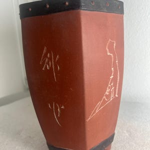 May include: A terracotta-colored, rectangular planter with a black rim and base. The planter has a decorative white etched design and Chinese characters. The planter is supported by three small feet.
