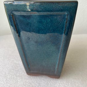 May include: A square, teal-colored ceramic planter with a glossy finish. The planter has a rectangular shape with a slightly tapered design. The interior is a contrasting brown color.