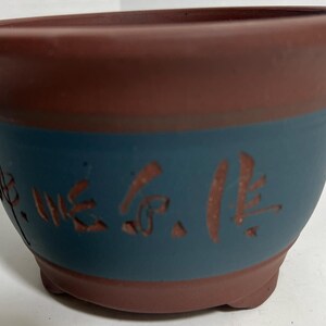 May include: A terracotta-colored ceramic pot with a blue band and brown Chinese characters around the middle. The pot has a rounded bottom and a wide opening.