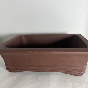 May include: A rectangular brown ceramic bonsai pot with a textured surface and a slightly raised rim. The pot has four small feet on the bottom.