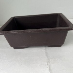 May include: A brown rectangular planter with a textured surface. The planter has a square shape and sits on four small feet. The sides are slightly sloped, and the rim has a subtle lip.
