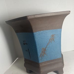 May include: A brown and blue ceramic planter with a geometric shape and three legs. The planter has a blue band with a gold design of a branch with leaves.