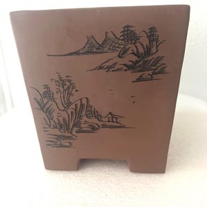 May include: A square, brown ceramic bonsai pot with a landscape design etched on the front. The design features mountains, trees, and a small building. The pot has a slightly tapered shape and small feet.