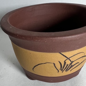 May include: A brown and tan ceramic planter with a black design. The planter has a wide, round opening and a slightly flared base.
