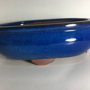May include: A blue ceramic bonsai pot with a rounded oval shape and a slightly flared rim. The pot has a glazed finish and rests on three small feet.