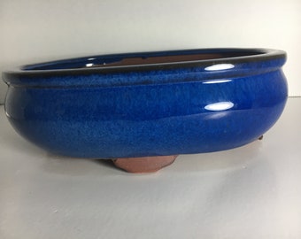 May include: A blue ceramic bonsai pot with a rounded oval shape and a slightly flared rim. The pot has a glazed finish and rests on three small feet.
