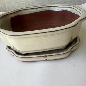 May include: Cream-colored ceramic planter with a rectangular shape and a matching saucer. The interior of the planter is a contrasting brown color. The planter has a slightly distressed, vintage appearance, suitable for plants.