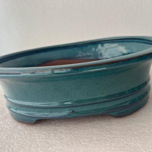 May include: An oval-shaped ceramic bonsai pot with a glossy, deep teal glaze. The exterior features a smooth, curved wall with a subtle horizontal ridge near the base. The interior reveals a natural, unglazed terracotta-colored clay surface.