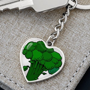 Puede incluir: Un llavero plateado en forma de corazón con un diseño de brócoli verde. La ilustración del brócoli presenta diferentes tonos de verde. El llavero está unido a una cadena plateada y a una llave.