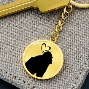 May include: A gold-colored keychain with a round pendant featuring a black silhouette of a dog with a heart above its head. The keychain has a gold chain and a gold key attached. The background is a textured, neutral-colored fabric.