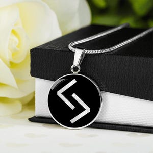 Può includere: Collana color argento con pendente rotondo. Il pendente ha uno sfondo nero con un simbolo runico bianco stilizzato. Presentata su una scatola nera, con un fiore bianco sullo sfondo.