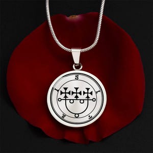 Op de afbeelding: Een zilverkleurige hangerketting met een ronde hanger met een zwart-wit sigil-ontwerp. De hanger hangt aan een zilveren ketting, tegen een donkerrode achtergrond.