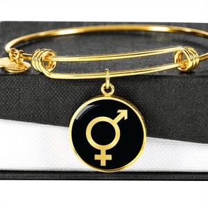 Puede incluir: Un brazalete de oro con un dije negro que presenta un símbolo de género dorado.