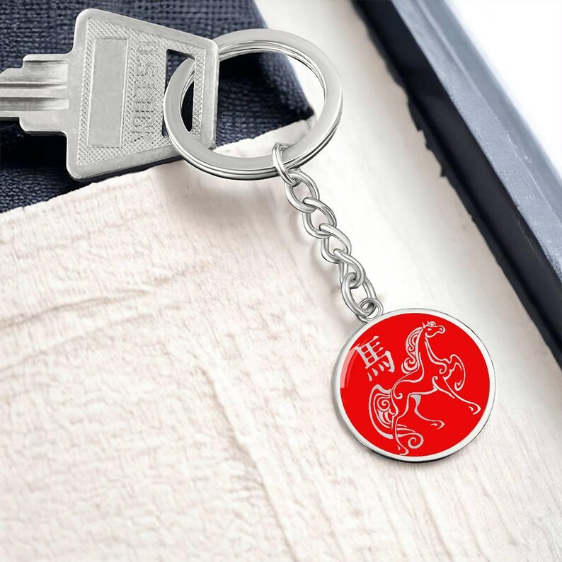 May include: A silver keyring with a round, red pendant. The pendant has a white horse illustration and Chinese characters. The keyring is attached to a key and a silver chain. The pendant has a silver border.