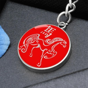 May include: A red circular keyring with a silver border. The keyring features a white illustration of a horse with wings and Chinese characters. The keyring is attached to a silver chain.