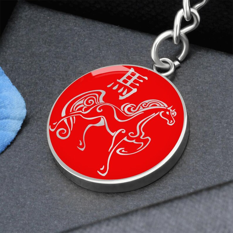May include: A red circular keyring with a silver border. The keyring features a white illustration of a horse with wings and Chinese characters. The keyring is attached to a silver chain.