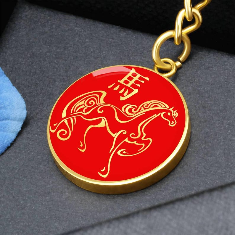 May include: A gold-coloured keyring with a red circular pendant. The pendant features a gold-coloured illustration of a horse with wings and Chinese characters. The keyring is attached to a gold-coloured chain.