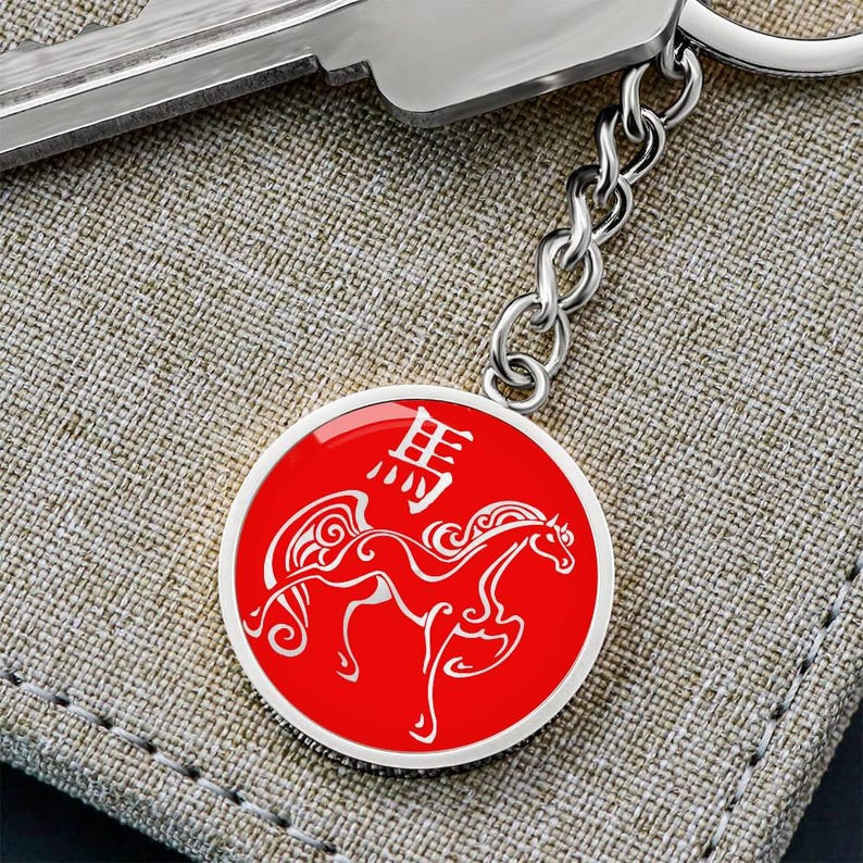 May include: A silver keyring with a round, red pendant. The pendant displays a white horse illustration and Chinese characters. The keyring is on a textured, beige surface.