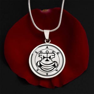 May include: A silver pendant necklace with a circular charm displaying a black and white sigil design. The pendant is suspended from a silver chain, set against a dark red background.