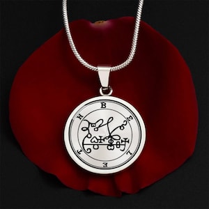 Op de afbeelding: Een zilverkleurige hangerketting met een ronde bedel met een zwart-wit sigil-ontwerp. De hanger hangt aan een dunne, zilveren ketting. De ketting ligt op een donkerrode ondergrond.