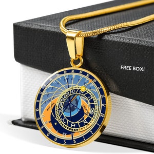 Pode incluir: Um colar de ouro com um pingente redondo que apresenta um mostrador de relógio astronômico azul e dourado com algarismos romanos e símbolos do zodíaco. O pingente está em uma caixa preta com as palavras "FREE BOX!" impressas nela.
