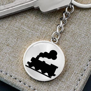 May include: A silver keychain with a round metal charm featuring a black silhouette of a train engine with smoke coming from the chimney.