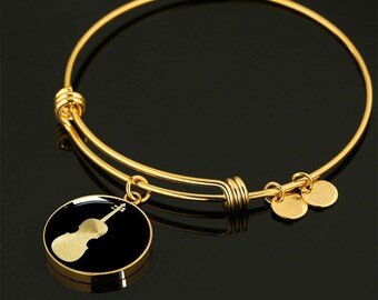 May include: A gold expandable bangle bracelet with a round charm featuring a black silhouette of a violin. The charm has a gold border and a shiny finish.