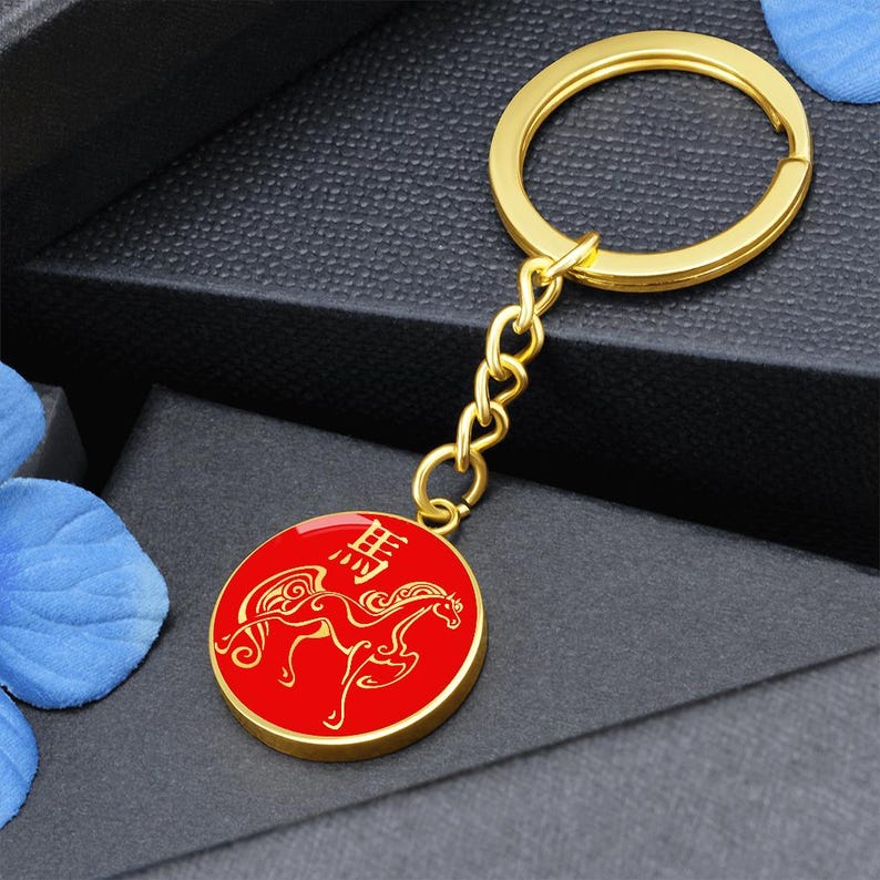 May include: A gold-coloured keyring with a red circular pendant. The pendant features a gold-coloured horse design and Chinese characters. The keyring has a gold ring and chain.