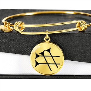 Może przedstawiać: Złota bransoletka typu bangle z okrągłym wisiorkiem przedstawiającym czarny symbol. Wisiorek jest przymocowany do bransoletki złotym łańcuszkiem.